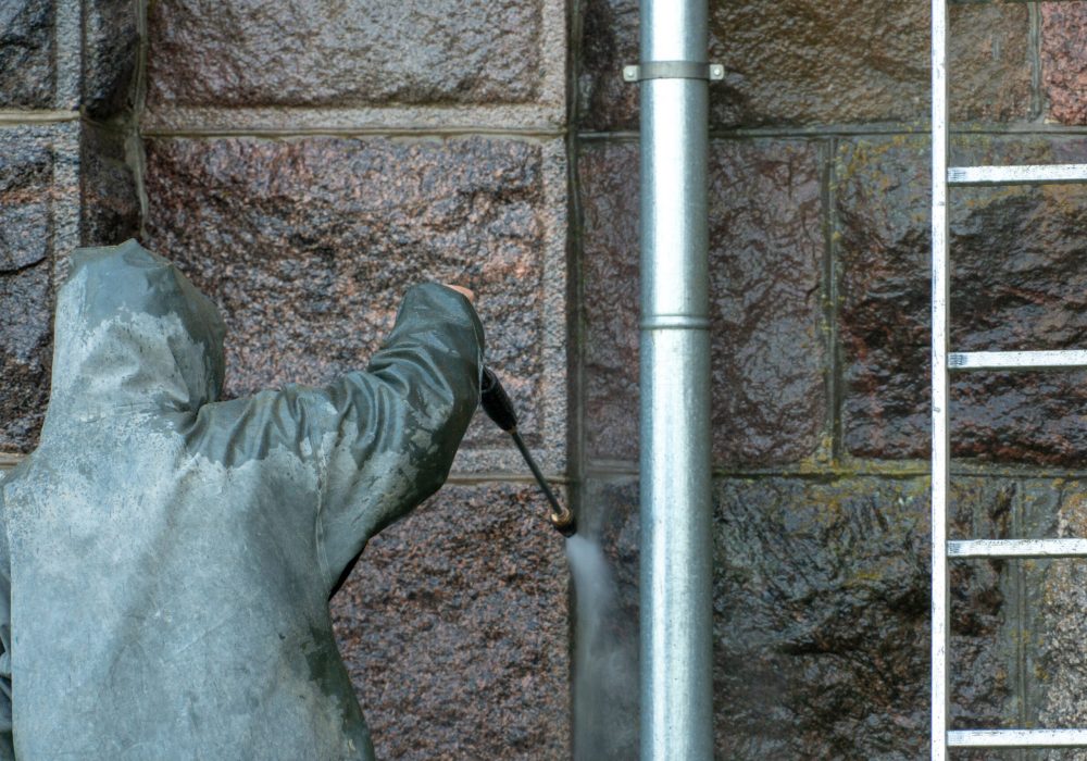 Man standing on a stepladder washes the walls of the building with a jet of water under pressure. Chemical protective suit. Decontamination of building from coronavirus. Security measures from covid19